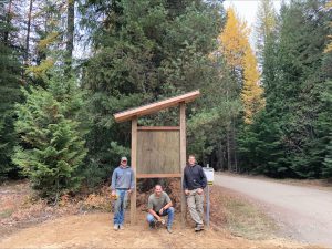 Three men sit in front of wood kiosk by narrow road and trees