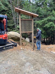 A man works on wood kiosk in front of trees with equipment.